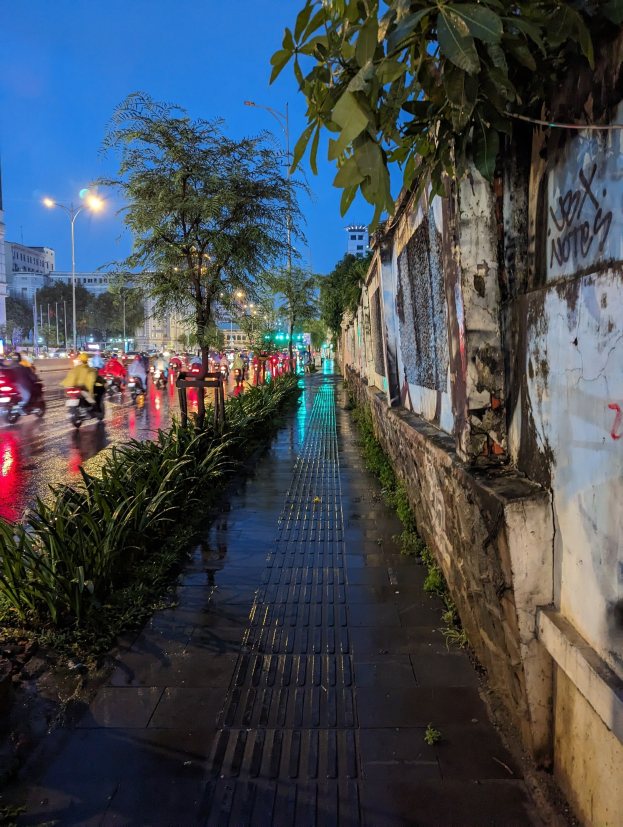 Stadtstraße bei Nacht mit Graffiti an der Wand, beleuchtet von Straßenlaternen, Fahrzeuge fahren, Bäume säumen die Seiten, Gebäude und sternenklarer Himmel im Hintergrund.