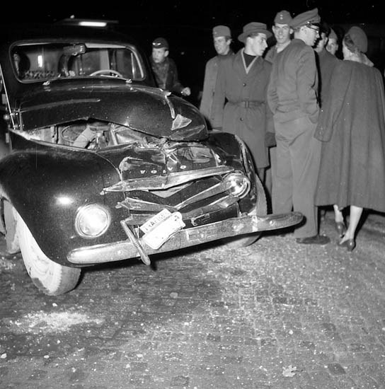 Black and white image of a severely damaged car on the roadside with a group of people inspecting it.
