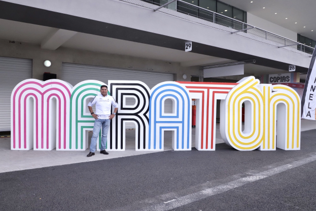 Ein Mann steht am Start eines Marathons vor einem großen "Marathon"-Schild, mit einem Banner rechts daneben und einem Gebäude mit Lampen, Geländern und Läden im Hintergrund.