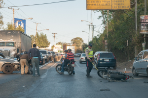 Gruppe von Menschen um ein verunglücktes Motorrad auf der Seite einer Straße mit mehreren Fahrzeugen, Bäumen, Masten, Lichtern, Schildern und Himmel im Hintergrund.