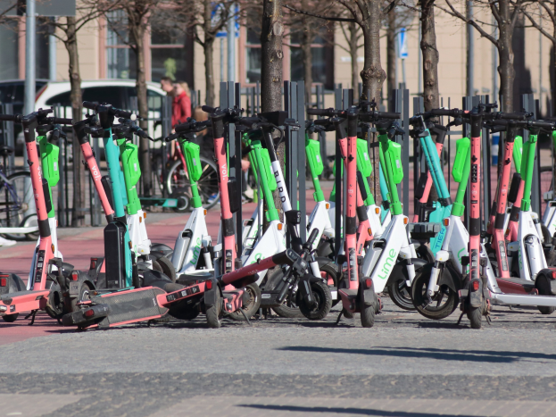 Eine Reihe von Elektro-Rollern, die an einer Straße geparkt sind, mit einer Person im Vordergrund und trees und Gebäude im Hintergrund.