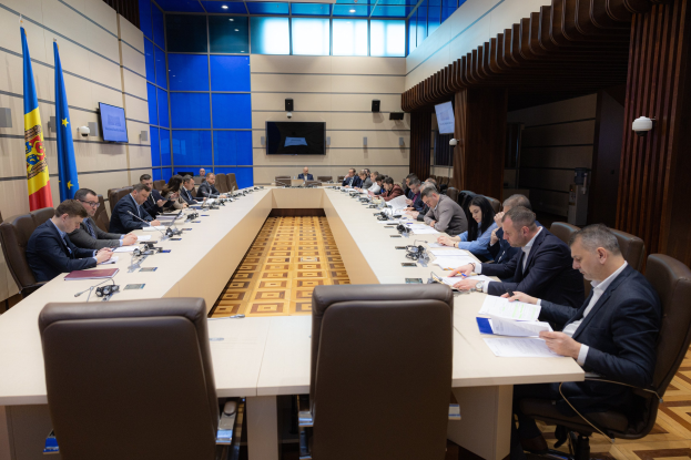 Gruppe von Menschen an einem langen Tisch mit Papieren, Büchern, Mikrofonen und anderen Gegenständen während einer Sitzung des Europäischen Parlaments in Brüssel, mit Fahnen auf der linken Seite und Bildschirmen und Lichtern im Hintergrund.