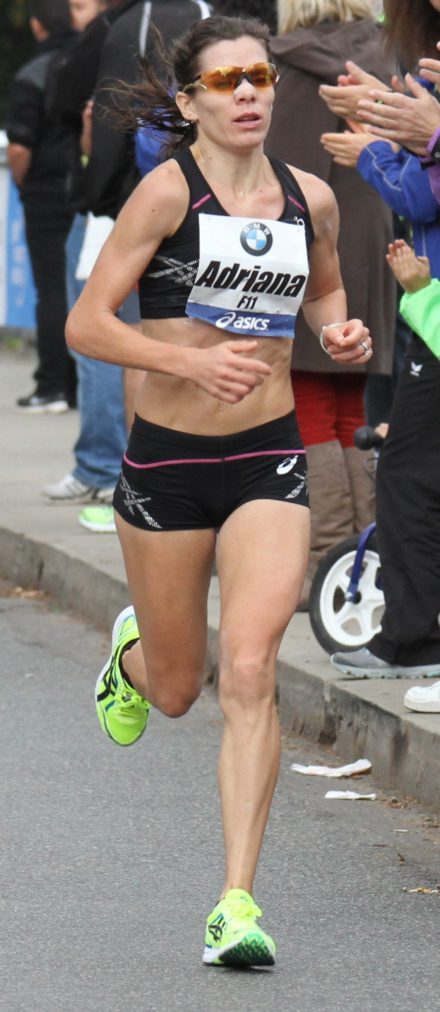 Eine Frau läuft bei einem Marathon in einem Sport-BH, Shorts und Goggles auf dem Kopf, mit einer Person auf dem Gehweg rechts daneben, ein paar Leute klatschen im Hintergrund und ein Rad ist im Bild zu sehen.