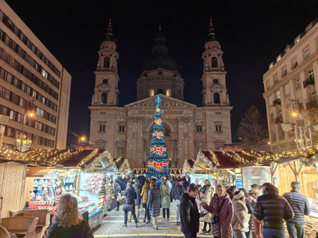 Ein belebter Weihnachtsmarkt mit geschmückten Ständen, festlicher Beleuchtung und Menschen vor einer Kirche bei Nacht, vor Gebäuden, Bäumen, Laternenmasten und einem sternenklaren Himmel.
