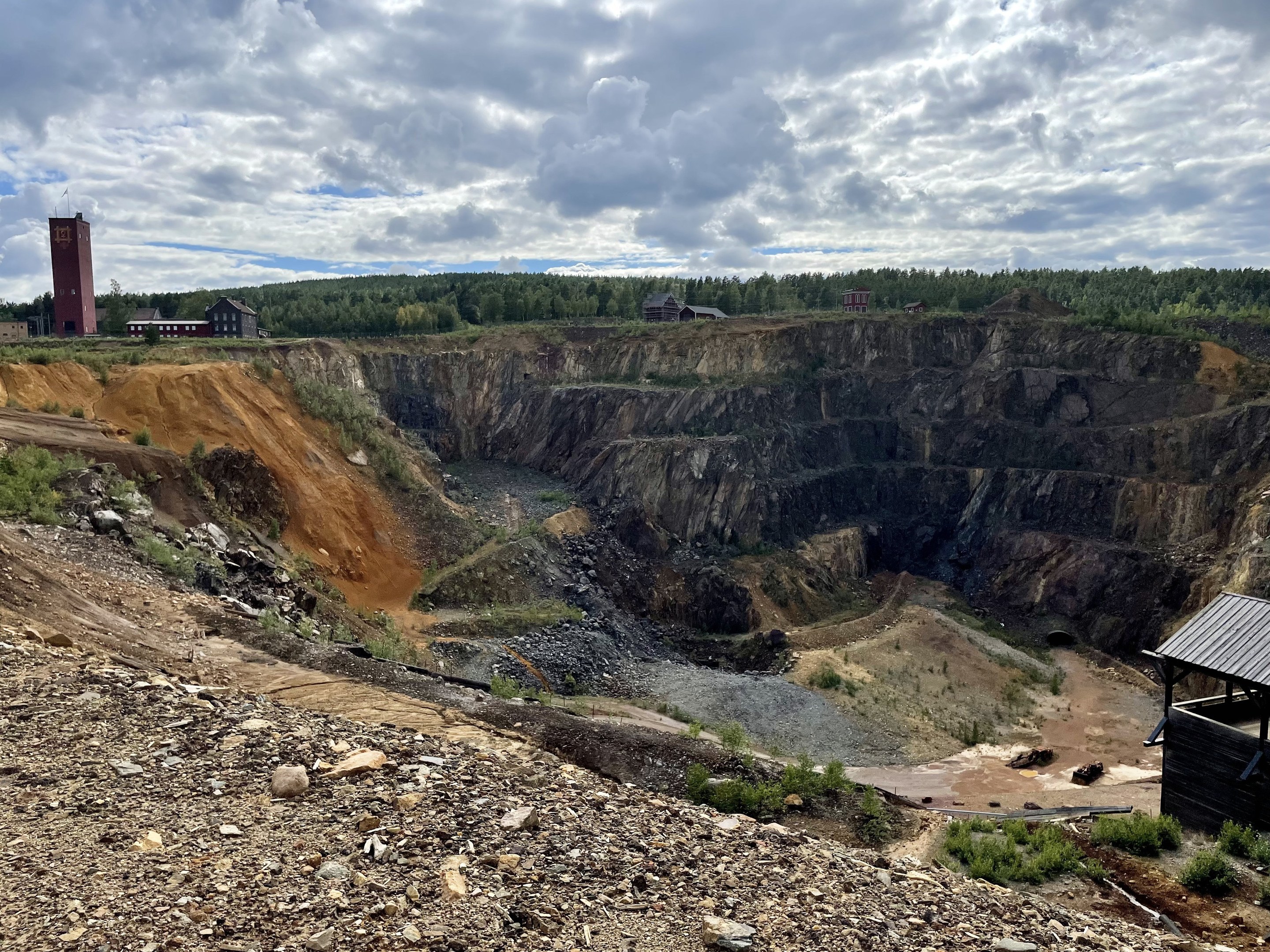 Großer Tagebau umgeben von Bäumen, Gebäuden und einem Schuppen, mit einem bewölkten Himmel und einem mit Steinen und Pflanzen bedeckten Boden.