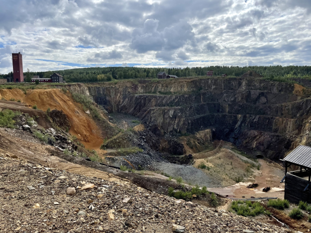 Großer Tagebau umgeben von Bäumen, Gebäuden und einem Schuppen, mit einem bewölkten Himmel und einem mit Steinen und Pflanzen bedeckten Boden.