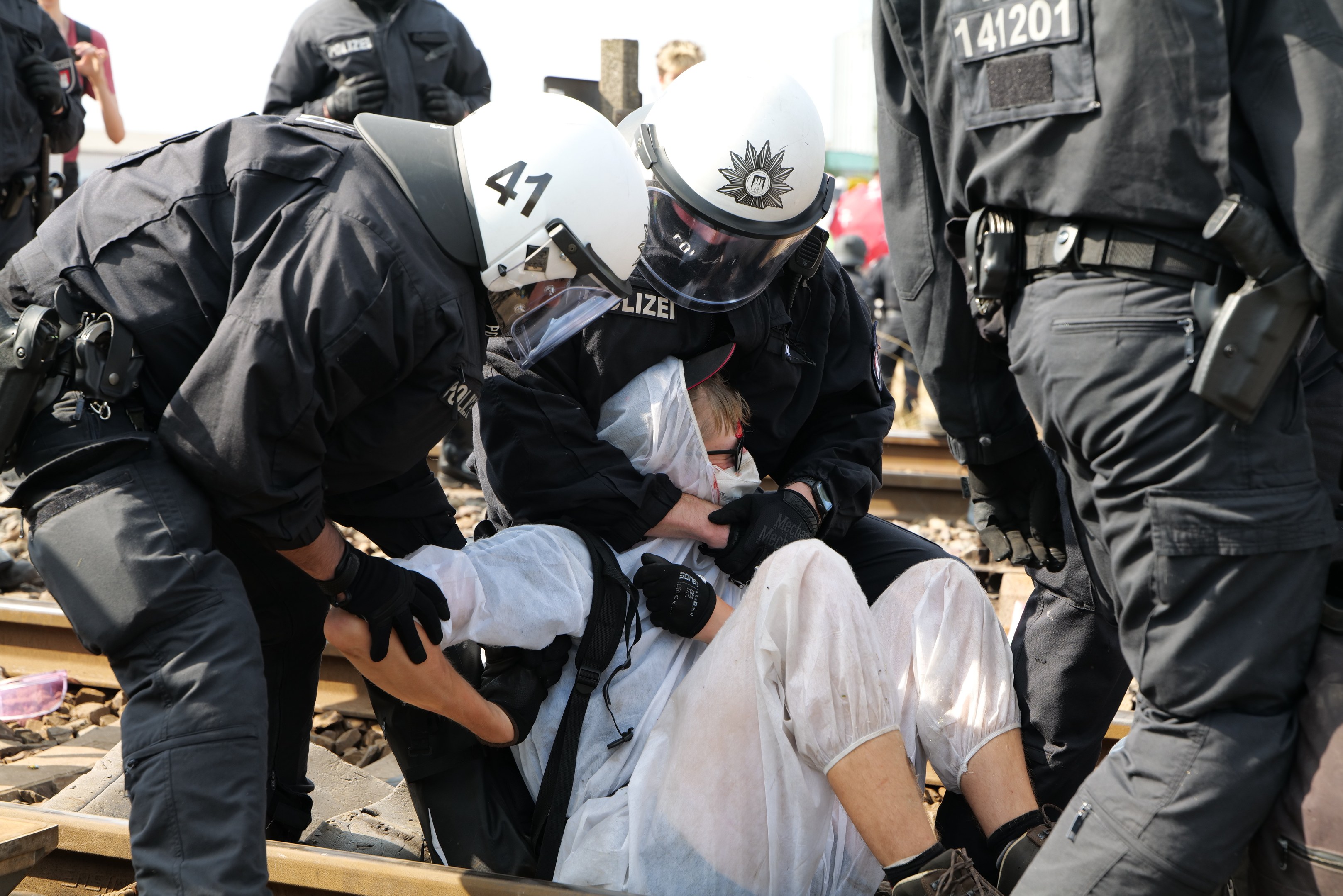Eine Gruppe von Polizisten in Einsatzausrüstung nimmt einen Mann in einem weißen Kleid auf einem Bahngleis fest, mit anderen im Hintergrund sichtbar.