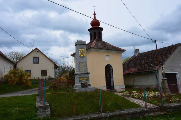 Kleine Kirche mit einem Glockenturm, identifiziert als die Kirche des Heiligen Grabes, umgeben von Häusern, Grünflächen, einem Zaun und einem bewölkten Himmel.
