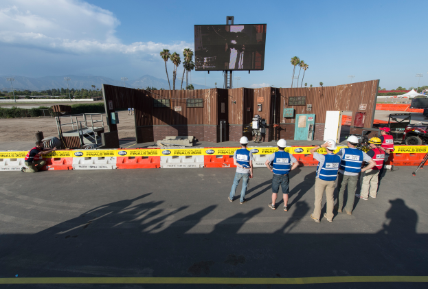 Eine Gruppe von Menschen in Helmen steht vor einem großen Bildschirm auf dem Las Vegas Motor Speedway, umgeben von Absperrungen, Fahrzeugen, Hütten, Bäumen, Lichtmasten und Bergen mit einem bewölkten Himmel im Hintergrund.