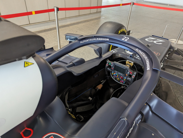 Cockpit eines Formel-E-Autos in einer Ausstellungshalle mit einem Lenkrad im Vordergrund, einer Wand im Hintergrund und dem Auto umgeben von Pfosten mit Bändern.