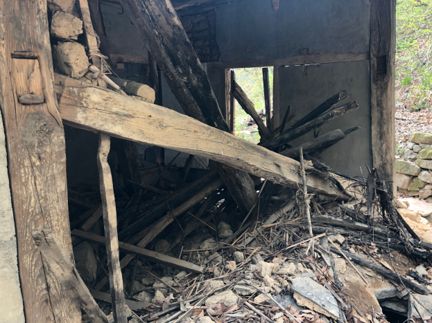 Ein altes Gebäude umgeben von Trümmern, einschließlich Holzstöcken, Steinen und trockenen Blättern, mit Bäumen im Hintergrund, scheint im Begriff zu sein, abgerissen zu werden.