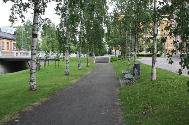 Ein Park mit einem baumgesäumten Weg, Bänken, Gras, einer Brücke, Gebäuden, Treppen, Laternen und Himmel.