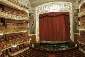 Großer Zuschauerraum mit roter Gardine, Sitzplätzen, Geländern, Beleuchtung, Wandskulpturen und Bühnenobjekten, die ein Konzertsaal vermuten lassen.