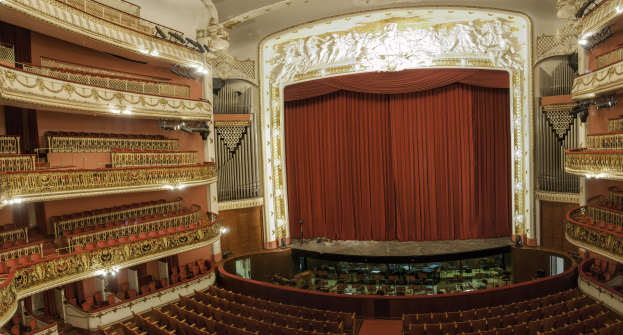 Großer Zuschauerraum mit roter Gardine, Sitzplätzen, Geländern, Beleuchtung, Wandskulpturen und Bühnenobjekten, die ein Konzertsaal vermuten lassen.