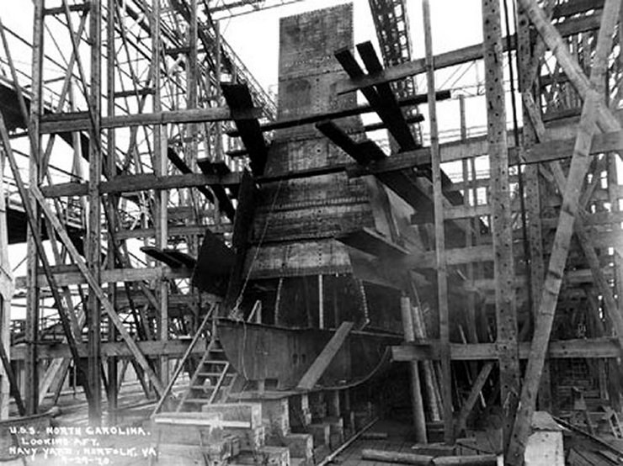 Schwarzes und weißes Foto eines Schiffs in einem Schiffbau, mit hölzernen Pfählen und Treppen im Vordergrund und Himmel im Hintergrund, beschriftet mit "Marinebewaffnung" in der linken unteren Ecke.