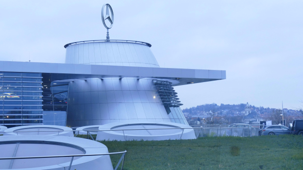 Das Mercedes-Benz Museum in Frankfurt, Deutschland, ein großes Gebäude mit einem Logo oben drauf, umgeben von Gras, Geländern, Fahrzeugen auf der Straße und Bäumen unter einem klaren blauen Himmel.