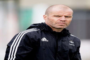 Mann mit Glatze in einem blauen Jackett mit weißen Streifen und weißem Kragen, der auf einem Fußballfeld steht und die Hände gefaltet hat.