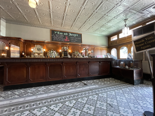 Ein großer Raum mit einer zentralen Bar, beleuchtet von an der Decke hängenden Lampen, mit Flaschen auf einem Tisch, einer Tafel mit Text an der rechten Seite, im Tom and Jerry Pub in London.