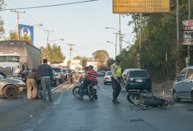 Eine Gruppe von Menschen umringt ein verunglücktes Motorrad am Straßenrand mit mehreren Fahrzeugen, darunter ein Lastwagen, und einer Hintergrundlandschaft aus Bäumen, Pfählen, Lampen und Schildern unter dem Himmel.