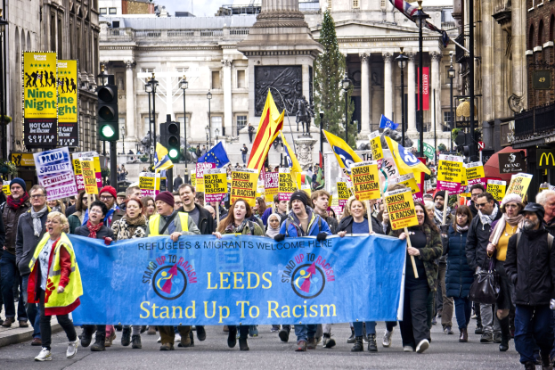 Eine Gruppe von Menschen marschiert mit einer 'Steh Auf gegen Rassismus'-Schleife die Straße entlang, mit Gebäuden, Bäumen, Laternen, Ampeln und einer Statue im Hintergrund unter einem bewölkten Himmel.