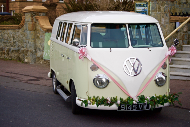 Ein weißer und pinker VW-Campervan mit floralen Verzierungen, der vor einer Steinwand geparkt ist, neben einer Treppe und einem Gebäude im Hintergrund.