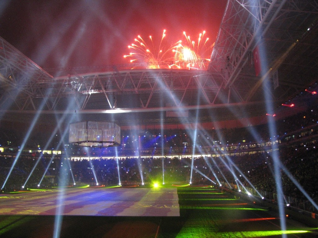 Das Olympische Stadion in London bei Nacht, erleuchtet von hellen Lichtern und Feuerwerken, mit einem großen Bildschirm im Vordergrund und einer Menge drinnen.