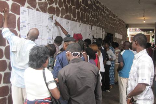 Eine Gruppe von Menschen steht vor einer Wand mit aufgeklebten Papieren, beleuchtet von Deckenlampen, wobei einige Personen Taschen tragen, die darauf hindeuten, dass sie wählen.