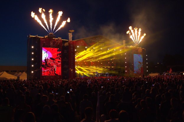 Eine große Menge bei einem Musikfestival schaut auf einer hell erleuchteten Bühne Performern zu, mit Zelten links und Feuerwerk am dunklen Himmel.