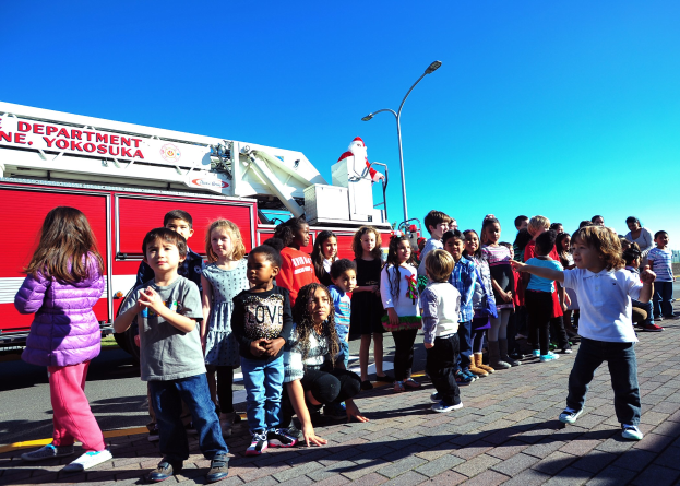 Kinder vor einem Feuerwehrfahrzeug, das für den Weihnachtsumzug der Yokosuka Fire Department beschriftet ist, auf einer Straße mit Laternenpfählen, Bäumen und einem klaren blauen Himmel.