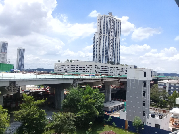 Luftaufnahme einer Stadtlandschaft mit Gebäuden, Bäumen, Gras, Straßen mit Fahrzeugen, einer Brücke, Strommasten und einem wolkenverhangenen Himmel, mit einem Baustellengelände für ein neues Wohnhochhaus.