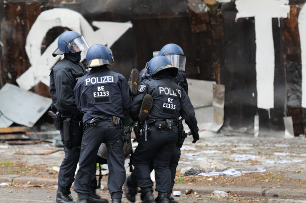 Polizisten in schwarzen Uniformen und blauen Helmen stehen vor einem mit Graffiti bedeckten Gebäude während einer Unruhewelle, mit verstreuten Papieren und Gegenständen auf dem Boden.
