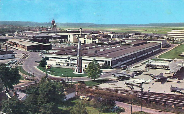 Alte Schwarz-Weiß-Airport-Szene mit geparkten Propellerflugzeugen, Terminalgebäuden, Bäumen, Straßen, Fahrzeugen, Versorgungsmasten, Grasflächen, Bahnschienen, fernen Türmen, Hügeln und einem klaren Himmel, mit gedrucktem Text entlang der unteren Kante.