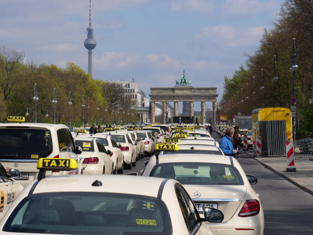 Eine belebte Straße in Berlin mit zahlreichen parkenden Taxis, Fußgängern auf dem Gehweg, Laternen, Bäumen, Gebäuden, einem fernen Bogen mit Statuen und Turm sowie einem bewölkten Himmel.