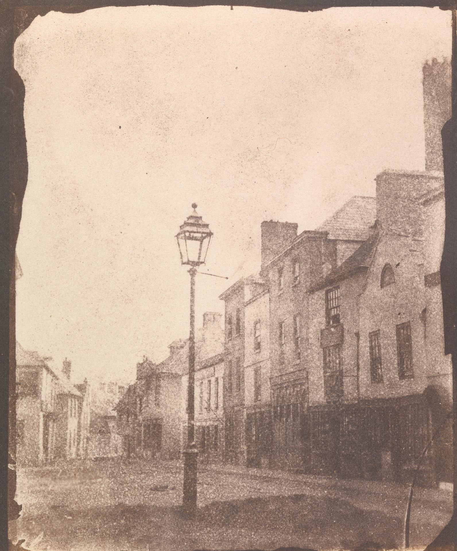 Alte Schwarz-Weiß-Fotografie einer Straßenlaterne in der Mitte einer Stadtstraße, flankiert von Gebäuden und einem klaren Himmel.