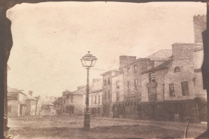 Alte Schwarz-Weiß-Fotografie einer Straßenlaterne in der Mitte einer Stadtstraße, flankiert von Gebäuden und einem klaren Himmel.