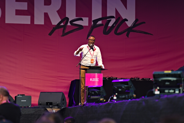 Ein Mann in einem weißen Hemd und braunen Hosen steht auf einer Bühne und hält ein Mikrofon, während er eine Menge anspricht. Im Hintergrund ist ein pinkes Banner mit der Aufschrift "Berlin as Fuck" zu sehen.