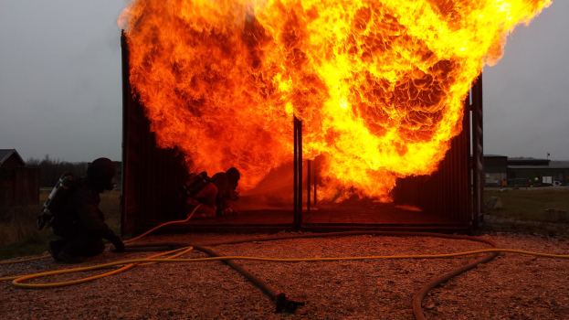 Feuerwehr löscht Gebäudebrand mit einer auf dem Boden liegenden Leitung, Schuppen und Bäume im Hintergrund bei klarem blauem Himmel.