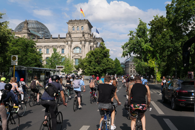 Eine Gruppe von Menschen, die auf Fahrrädern eine von Bäumen gesäumte Straße in Berlin, Deutschland, entlangfährt, mit einer Bushaltestelle auf der rechten Seite und einer Flagge auf einem nahegelegenen Gebäude.