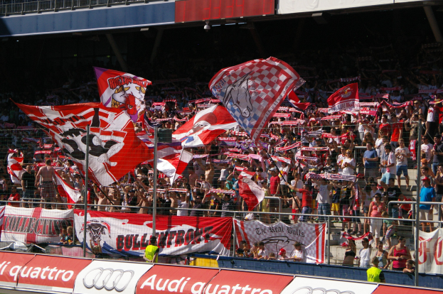 Eine große Menschenmenge in einem Stadion mit Fahnen, Bannern, einem Metallzaun, einer Anzeigetafel, einem Bildschirm und Deckenleuchten.