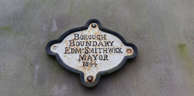 Metallplakette an einem Gebäude mit schwarzem Hintergrund und weißer fettgedruckter Schrift, die "Stadtteilgrenze Edm Smithwick Bürgermeister" liest.