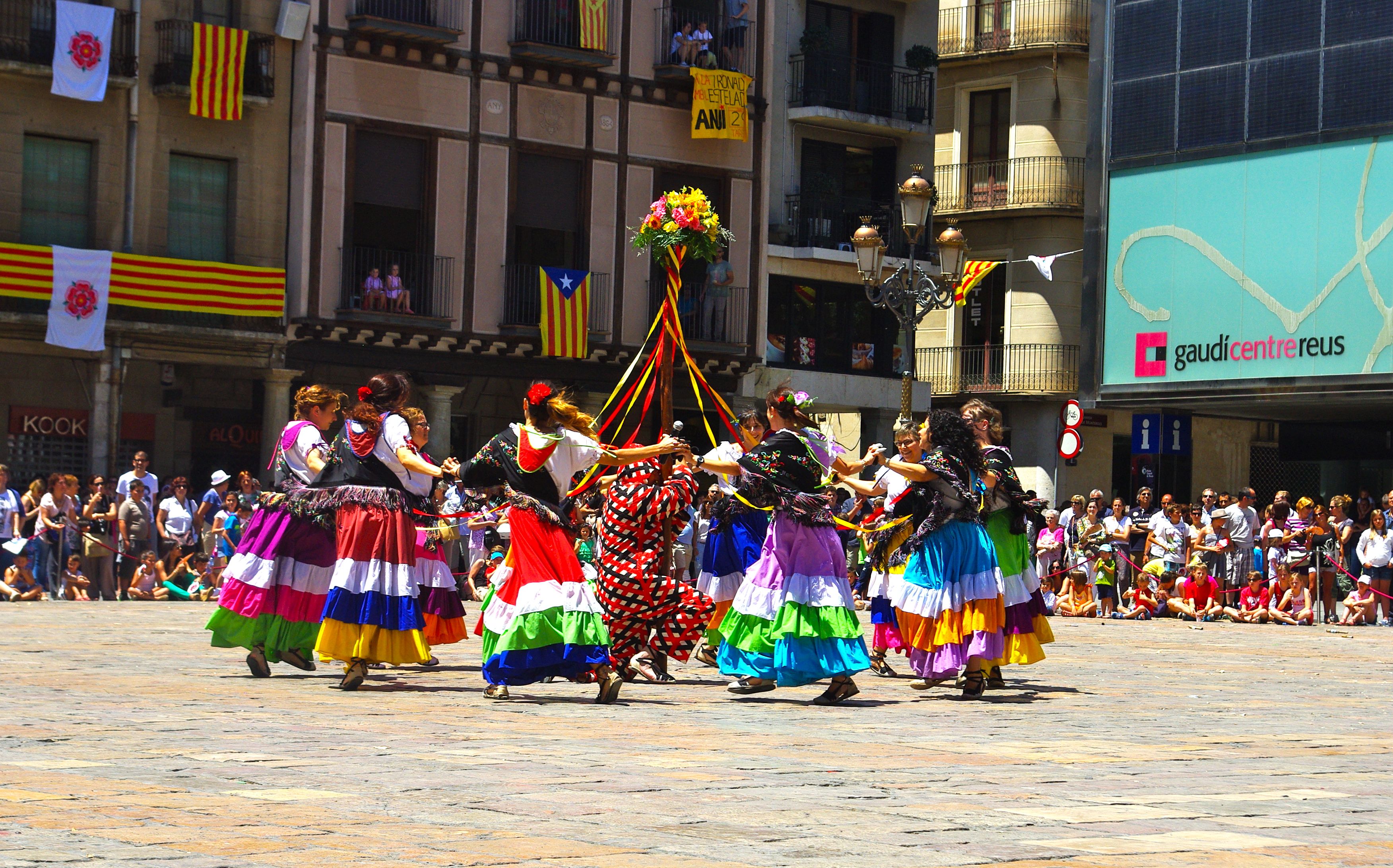 Eine Gruppe von Frauen in bunten Kleidern tanzt auf der Straße, umgeben von einer Menge Menschen, mit Gebäuden, Gelöndern, Balkonen, Flaggen, einem Laternenpfahl und einer Tafel mit Aufschrift im Hintergrund.