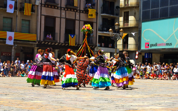 Eine Gruppe von Frauen in bunten Kleidern tanzt auf der Straße, umgeben von einer Menge Menschen, mit Gebäuden, Gelöndern, Balkonen, Flaggen, einem Laternenpfahl und einer Tafel mit Aufschrift im Hintergrund.