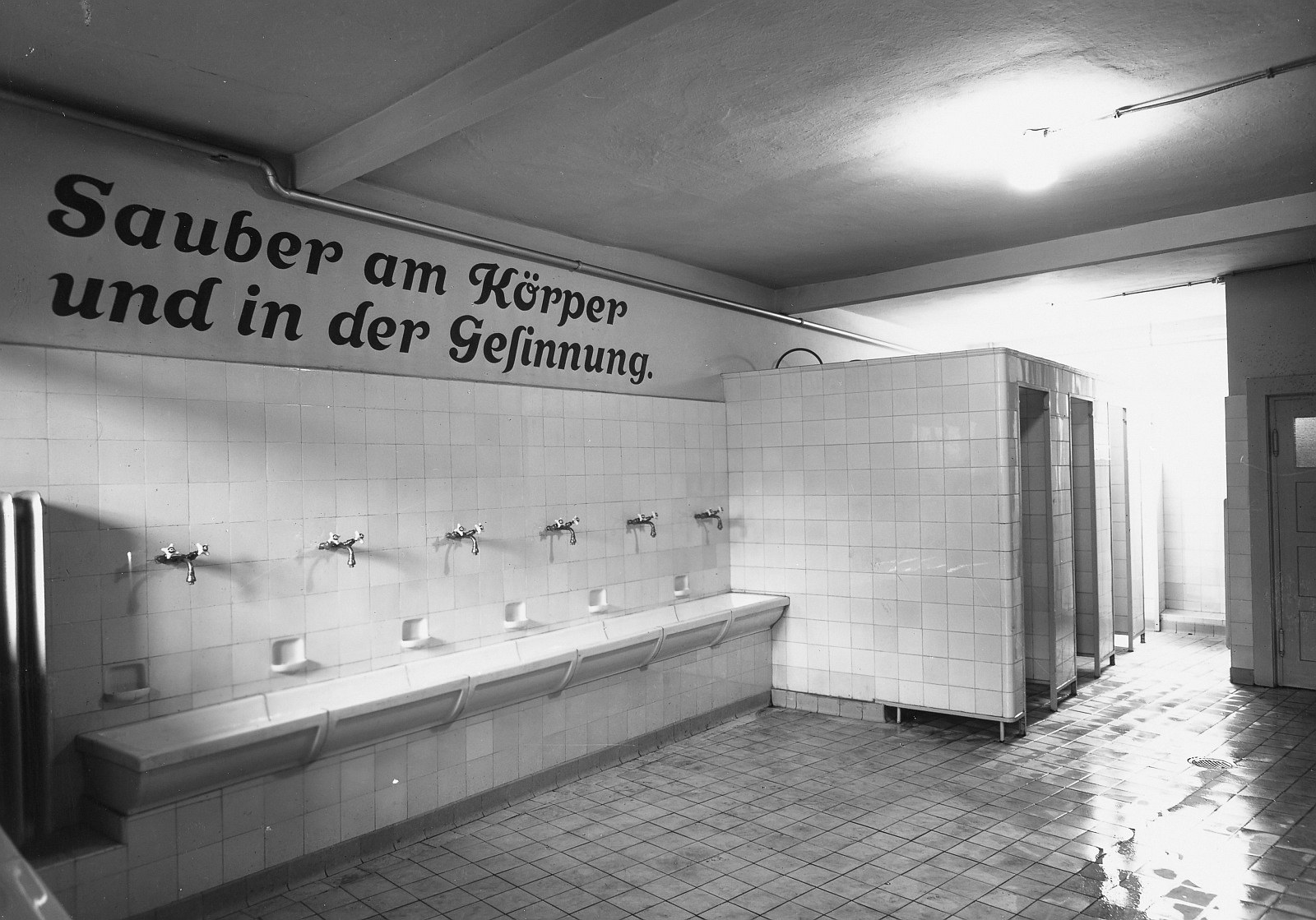 Schwarzes und weißes Foto einer öffentlichen Toilette mit Fliesenboden, einer Wand mit Text, mehreren Wasserhähnen, einer Tür und einer Deckenlampe.