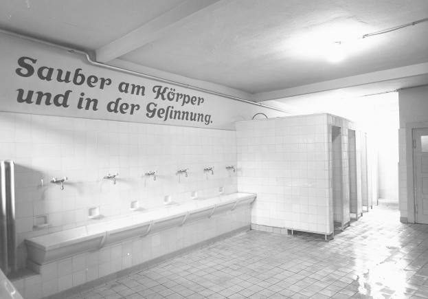 Schwarzes und weißes Foto einer öffentlichen Toilette mit Fliesenboden, einer Wand mit Text, mehreren Wasserhähnen, einer Tür und einer Deckenlampe.