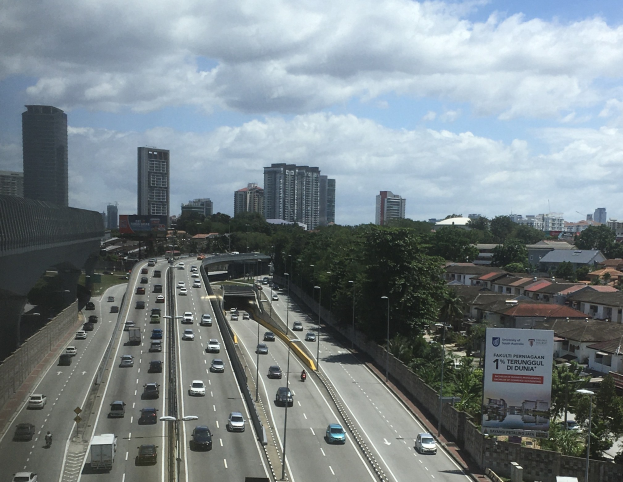 Eine belebte Autobahn mit hohen Gebäuden, einer Brücke auf der linken Seite, Laternenmasten, Bäumen und einem bewölkten Himmel im Hintergrund.