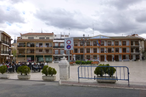 Ein belebter Stadtplatz mit Menschen, die sitzen und stehen, Topfpflanzen, Metallabsperrungen, ein Schild an einem Pfahl, Straßenlaternen mit Flaggen, umgebende Gebäude und ein bewölkter Himmel.