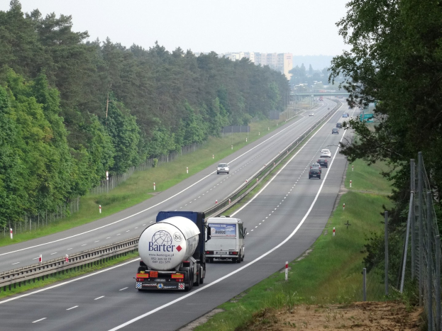 Ein großer LKW fährt auf einer von beiden Seiten mit Bäumen gesäumten Autobahn, mit einem Zaun und Gras auf der rechten Seite und Gebäuden im Hintergrund unter einem klaren blauen Himmel.