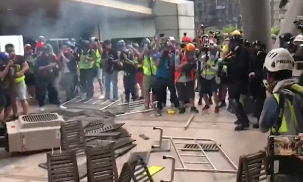 Menschen in Helmen und Jacken stehen vor einem rauchenden Gebäude, einige halten Kameras, mit verstreuten Metallobjekten und Stadtpfeilern im Hintergrund.