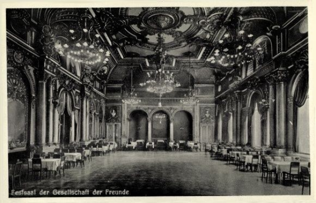 Schwarzes und weißes Foto eines großen Raums mit Tischen, Stühlen, Säulen, Bögen und Deckenleuchtern, beschriftet mit 'Festival der Gesellschaft der Freunde, Berlin' unten.