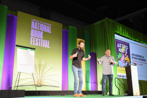 Zwei Männer stehen auf einer Bühne mit einem Podium und Mikrofon, vor einem großen Bildschirm und einem Banner mit der Aufschrift 'National Book Festival.'
